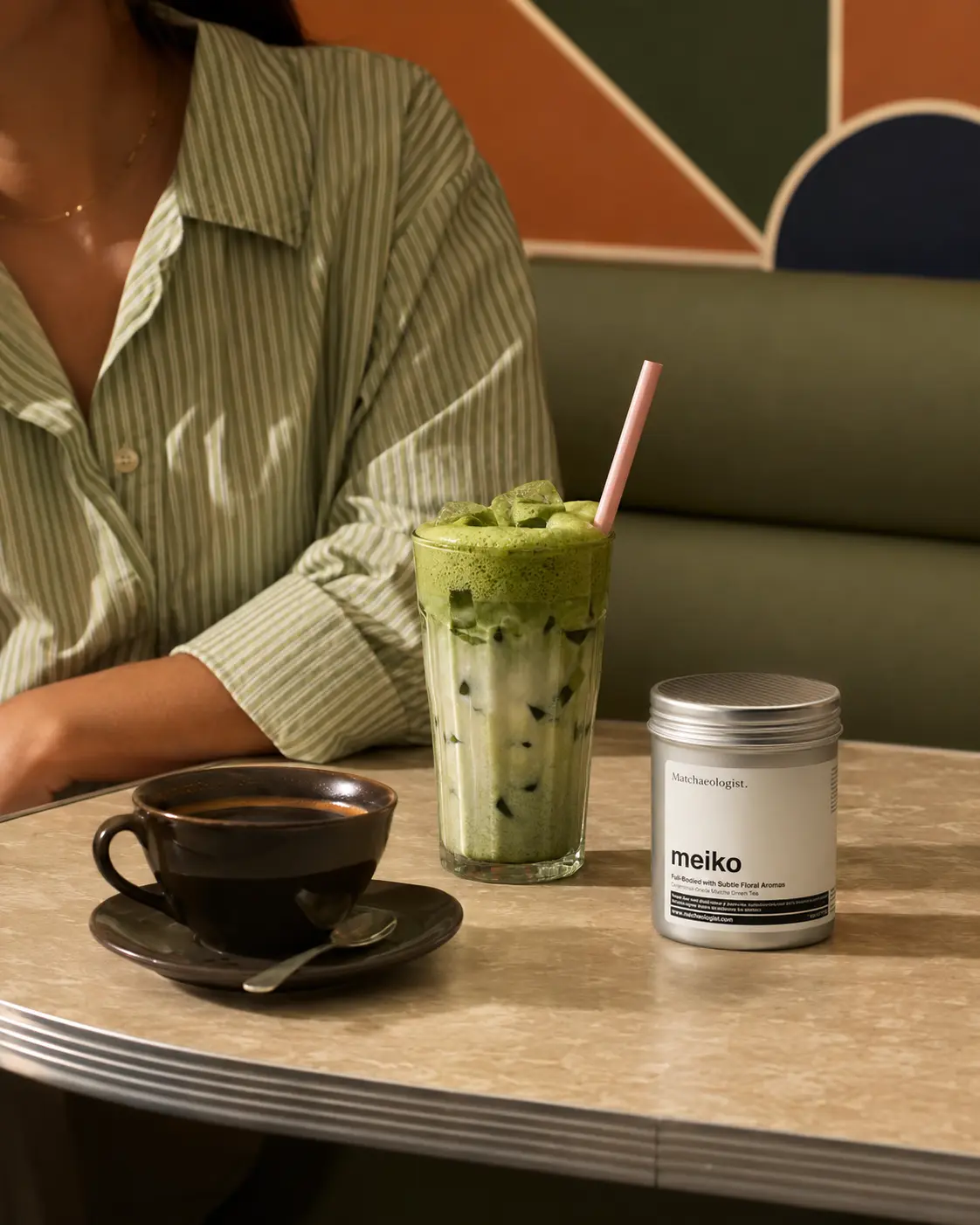 Meiko matcha tin beside an iced matcha and coffee in a warm retro café
